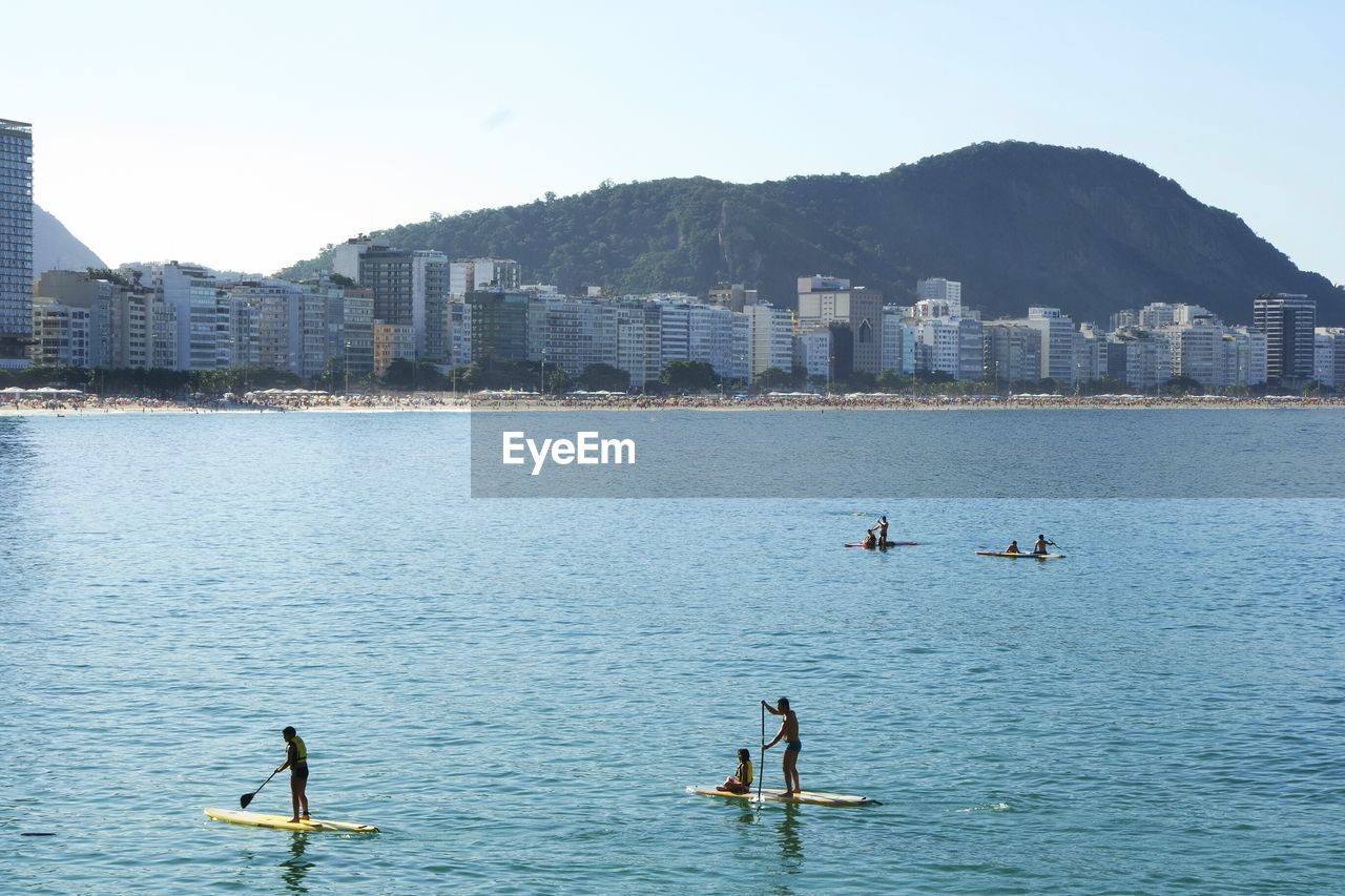 People paddleboarding in sea by city against sky