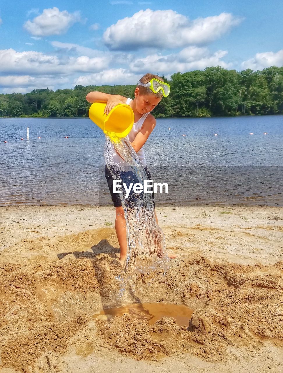 Boy pouring water on sand while standing against sea at beach