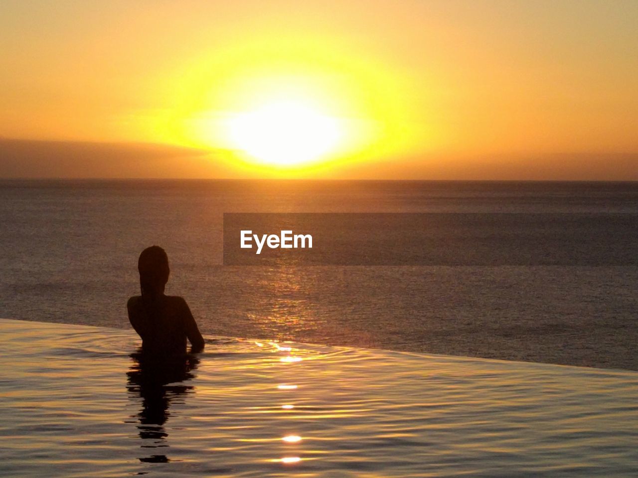 SILHOUETTE MAN SITTING AT SEA AGAINST SKY DURING SUNSET