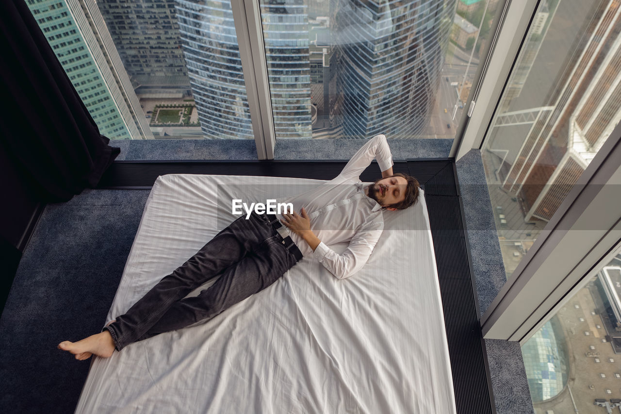 Businessman man in a white shirt is lying on a bed near a large panoramic window resting
