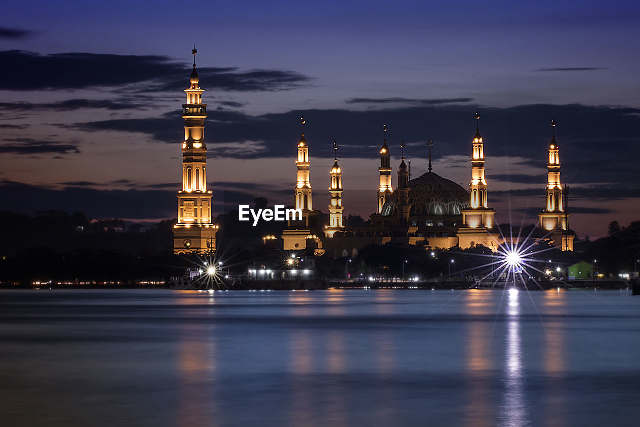 Illuminated mosque by river against sky at night