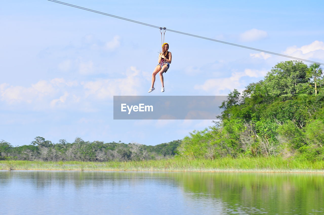 Low angle view of woman hanging from zip line over lake against sky