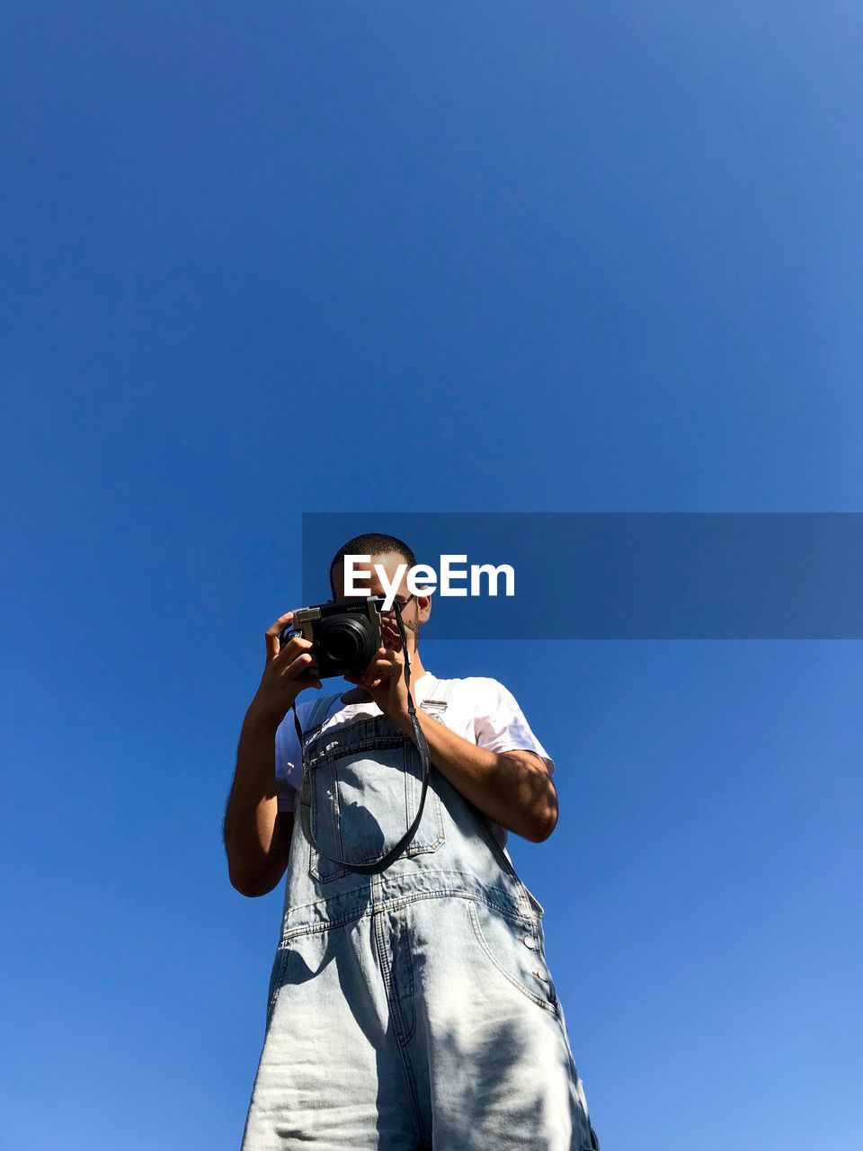 Low angle view of man photographing through camera while standing against clear blue sky
