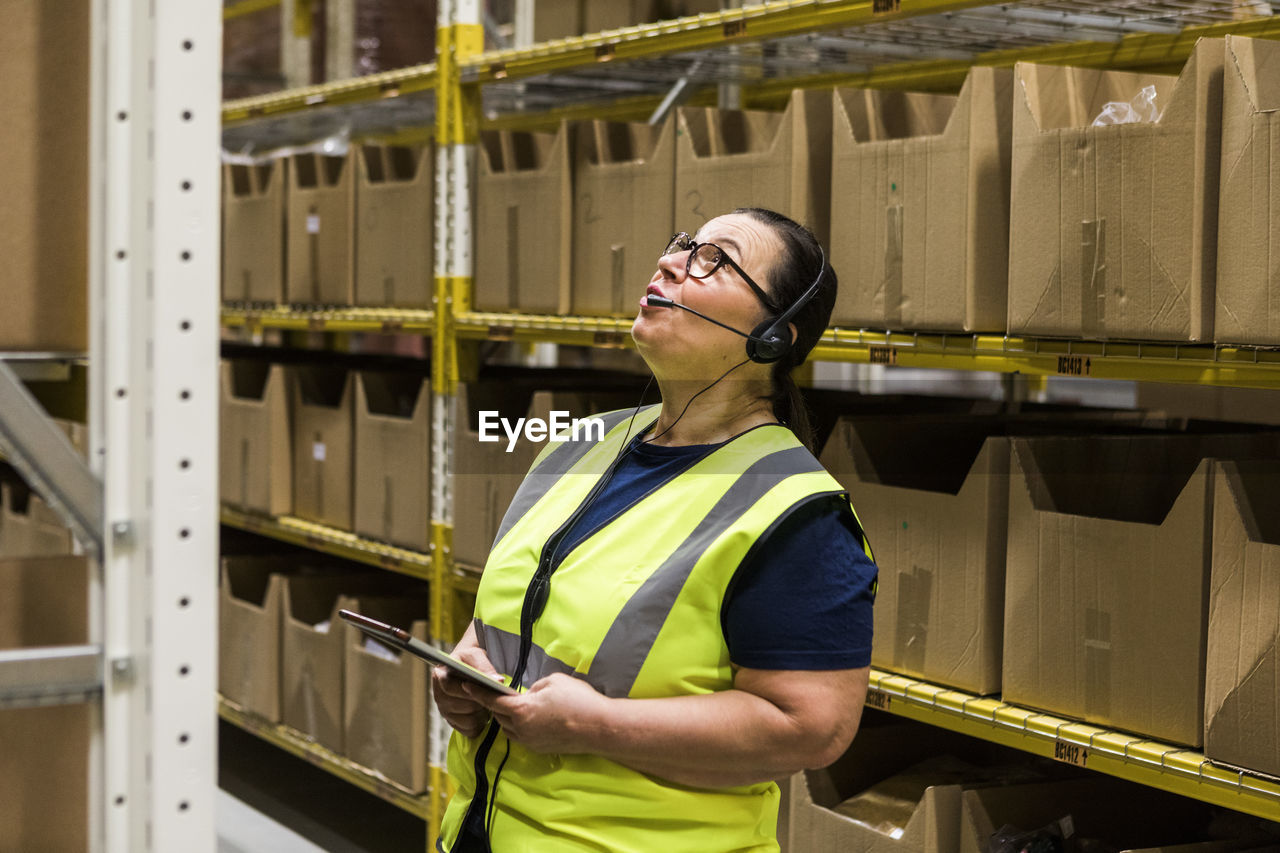 Mature female worker with digital tablet looking up while talking on headset against rack at distribution warehouse