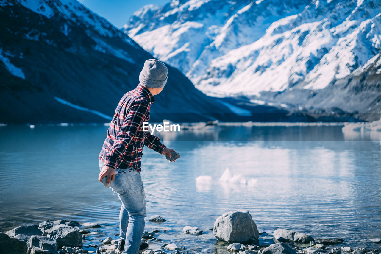 Man skipping rocks.man standing on rock by lake against snowcapped mountains during winter. 