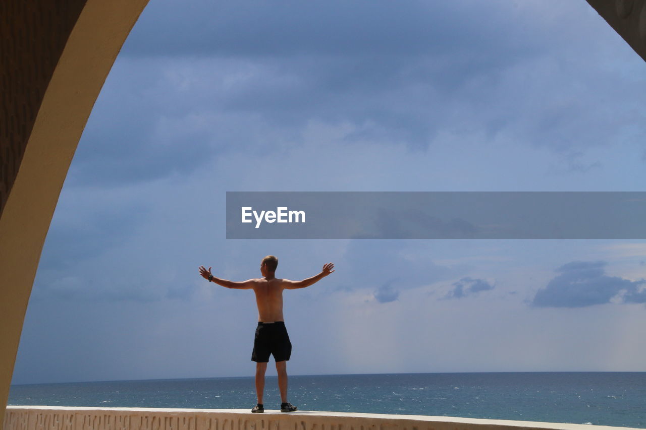 Rear view of shirtless man with arms outstretched standing on retaining wall by sea against sky