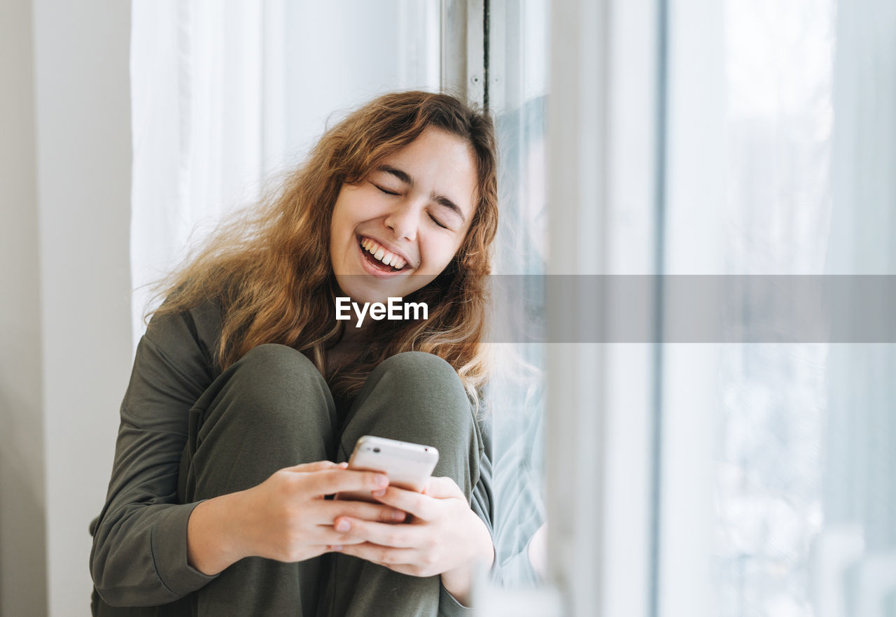 Beautiful smiling teenager girl with curly hair using mobile phone sitting on the window sill