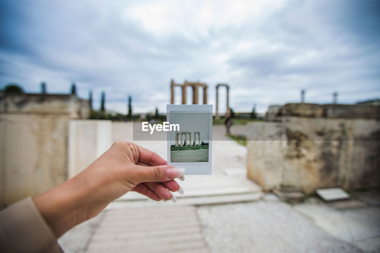 Cropped woman holding photograph at olympian zeus temple against cloudy sky
