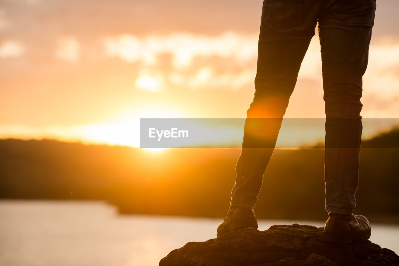 LOW SECTION OF MAN STANDING ON ROCK AGAINST ORANGE SKY