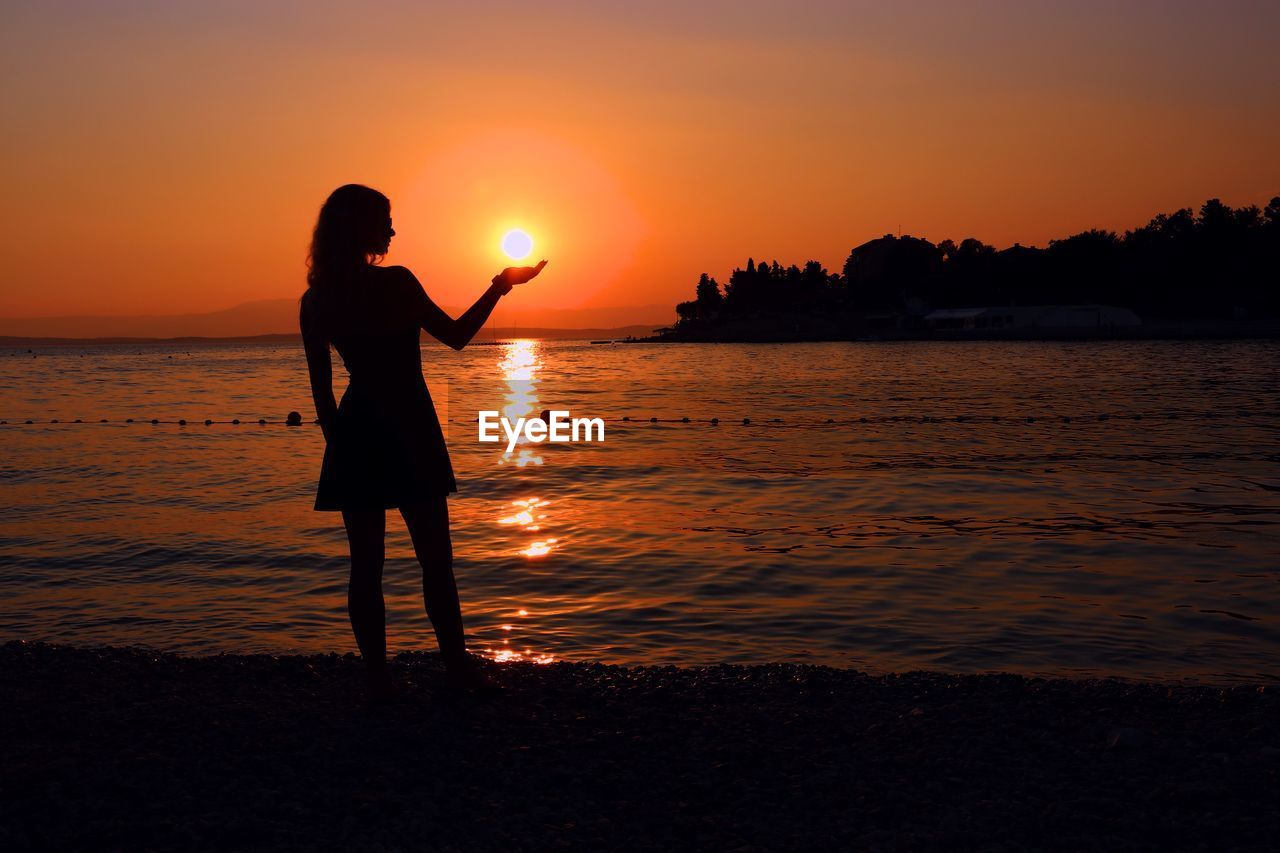 Optical illusion of silhouette woman holding sun while standing at beach against sky during sunset