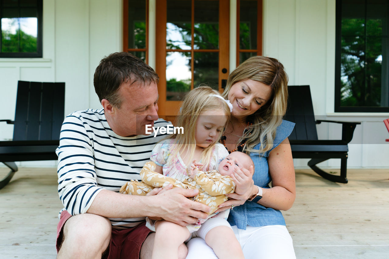 A young girl holds her newborn baby sister with her parents on a porch