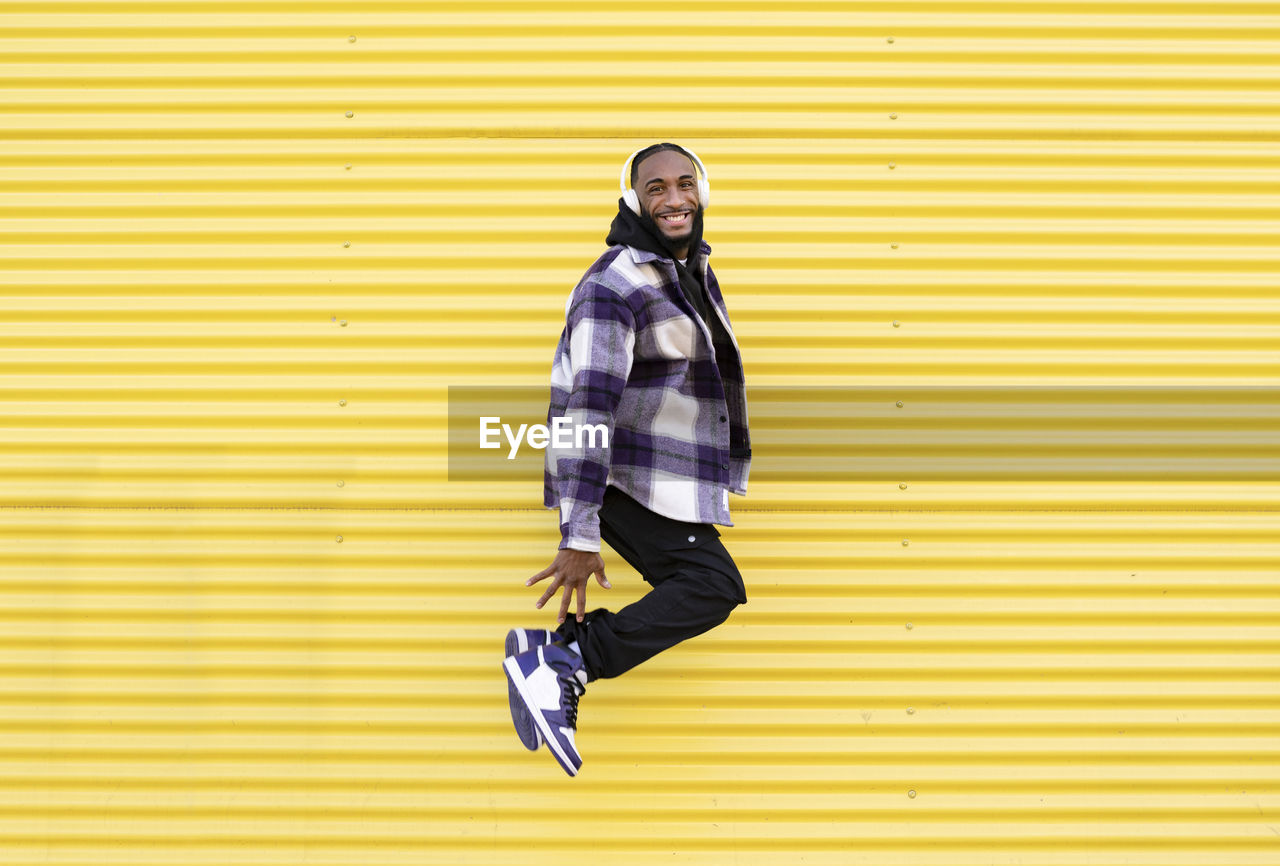 Happy young man in mid-air listening music through headphones against yellow corrugated iron