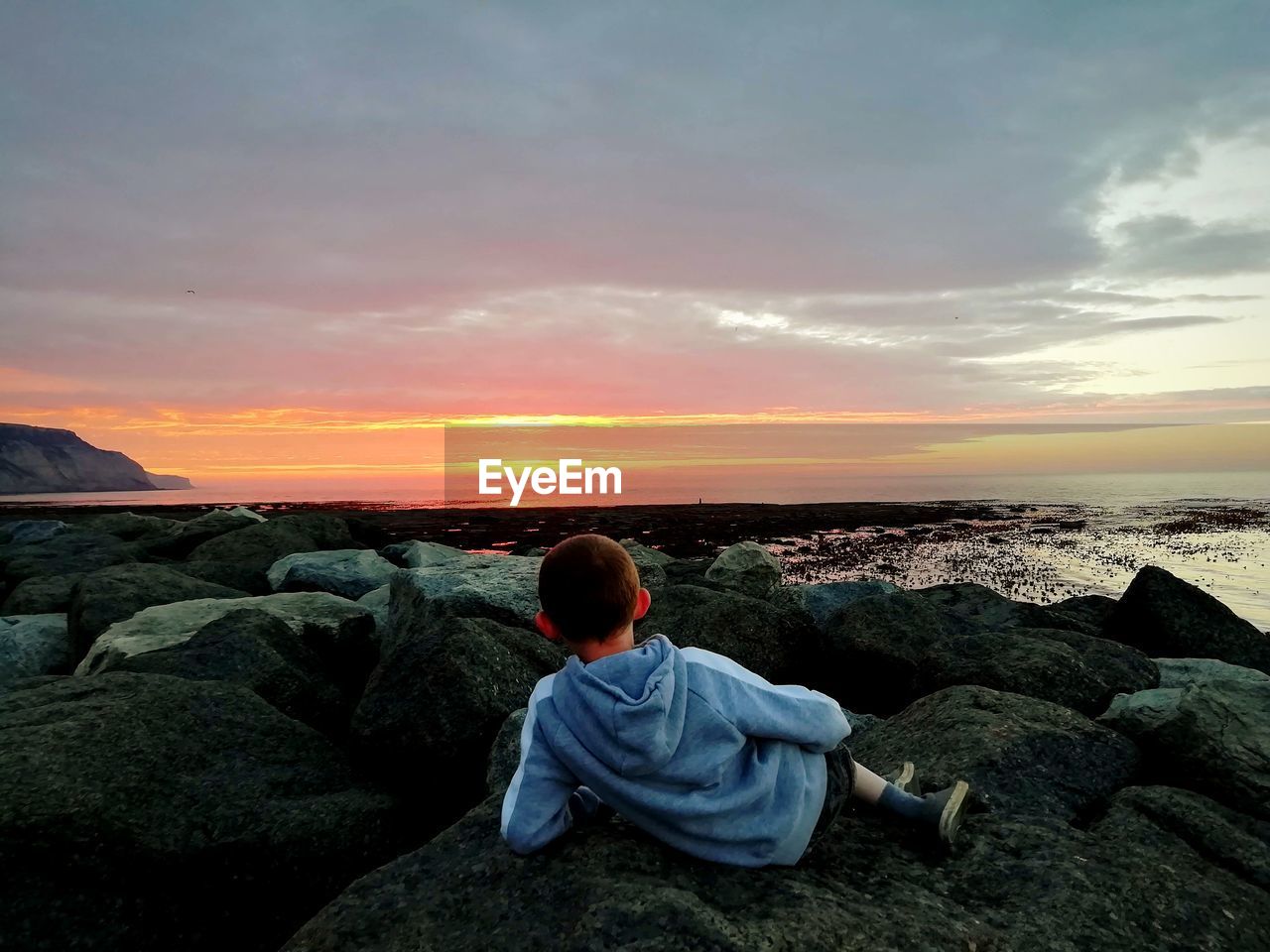 Rear view of boy sitting on rock by sea against sky