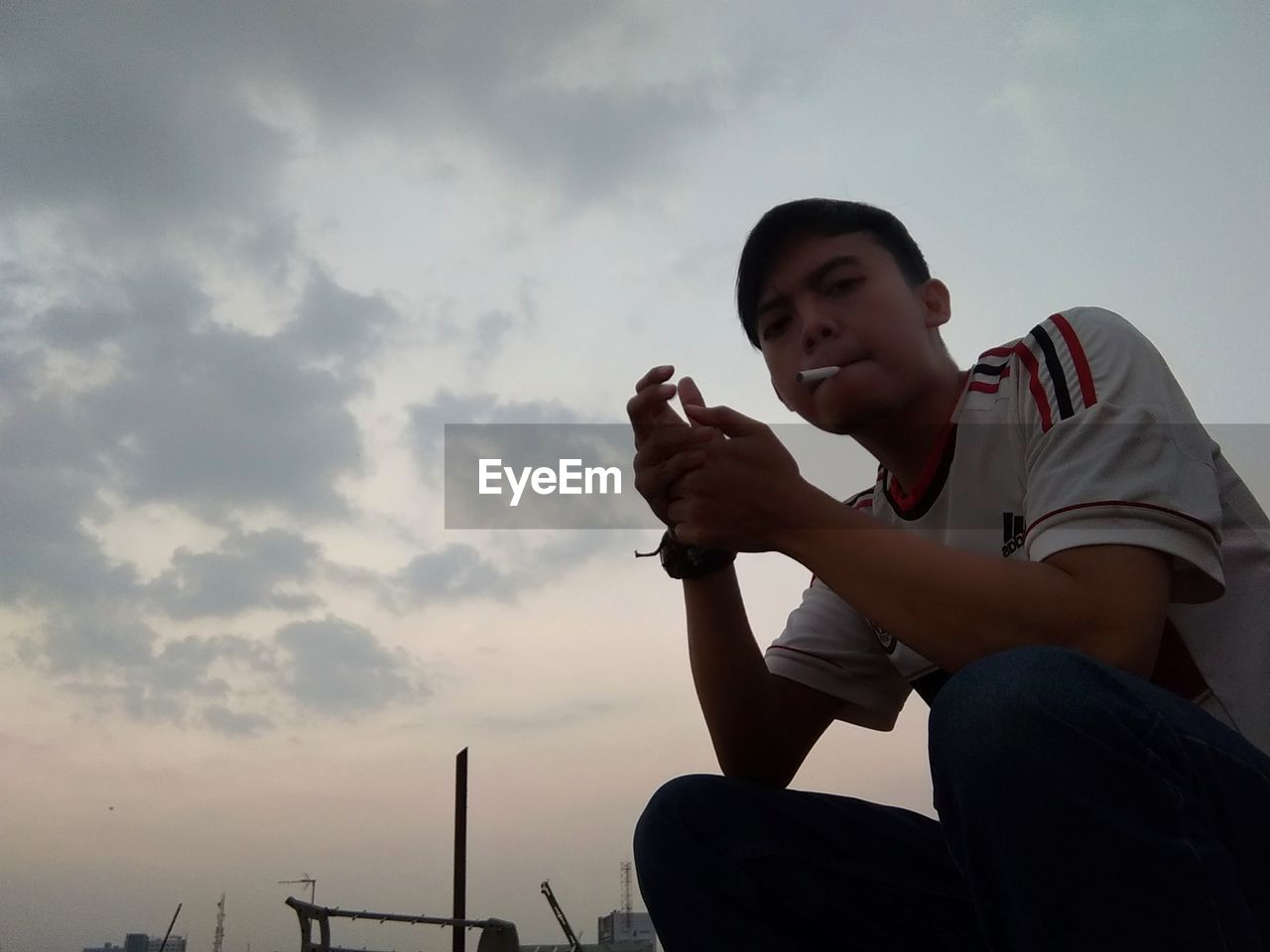 LOW ANGLE VIEW OF MAN SITTING ON CAMERA AGAINST SKY AGAINST CLOUDY