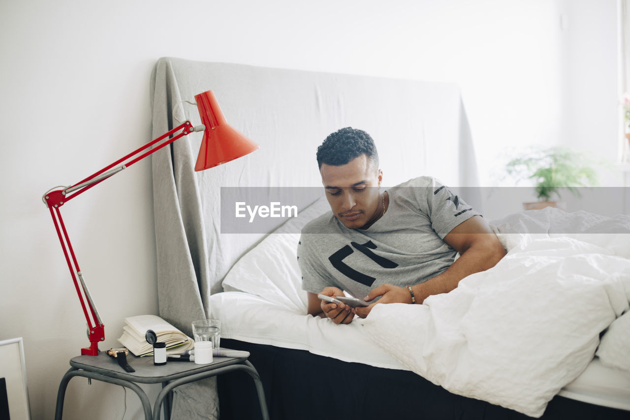 Mid adult man holding glaucometer and mobile phone while lying on bed by side table