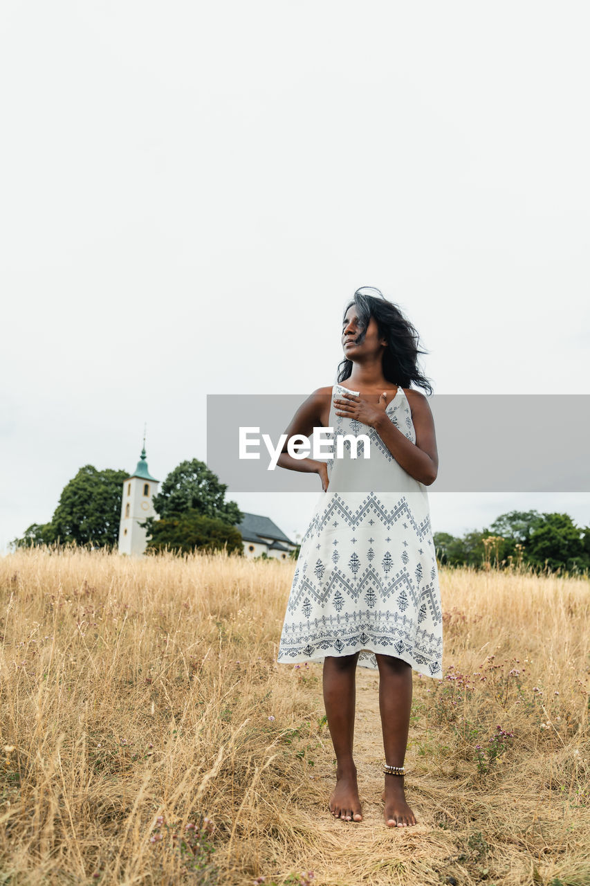 Barefoot adult ethnic female in sundress touching chest while looking away on pathway against chapel under light sky