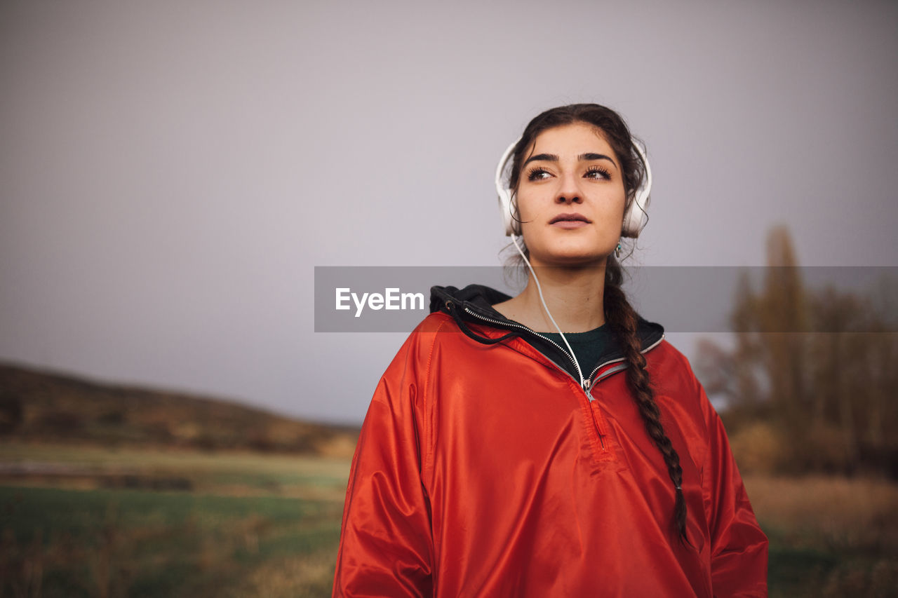 Beautiful young woman wearing headphones while standing on field against sky