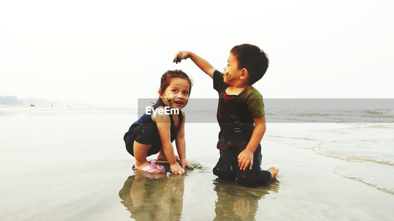 Siblings playing with sand at beach against clear sky