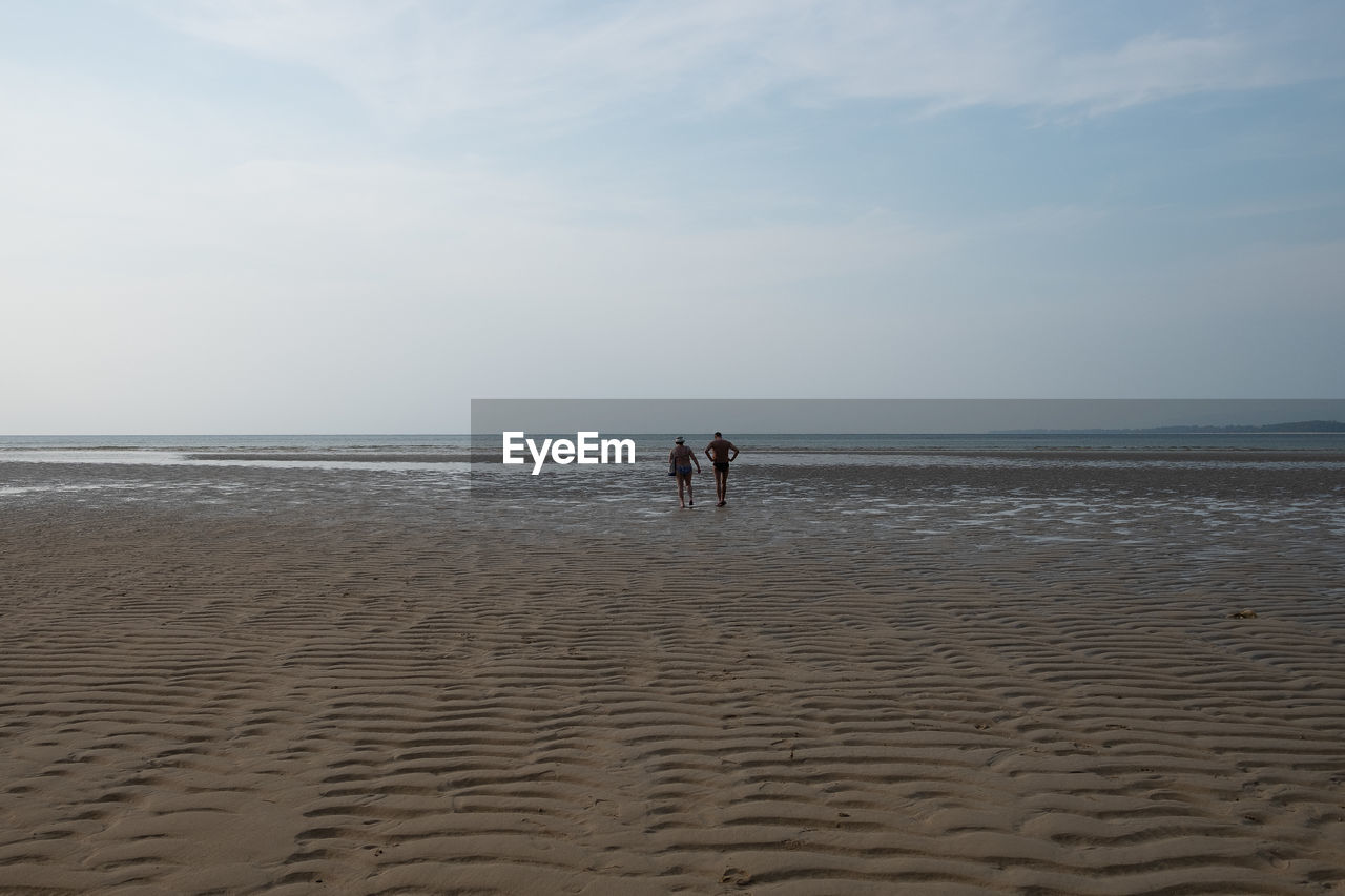 Rear view of man and woman walking towards shore at beach against sky