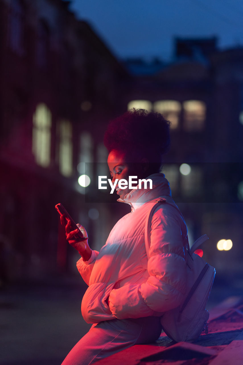 Young smiling african american woman with smartphone standing outdoors at night