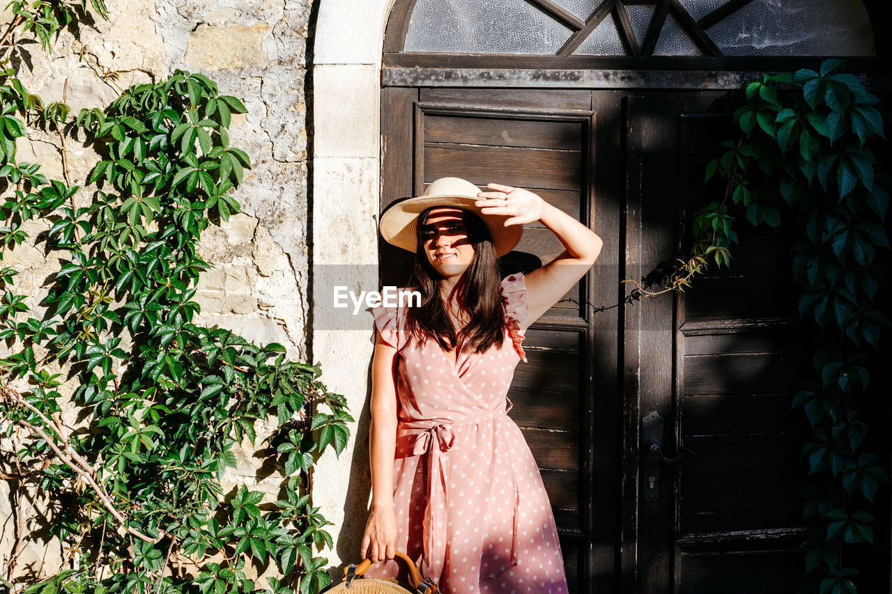 Beautiful young woman in pink dress standing in front of picturesque old house, door, light, shadow.