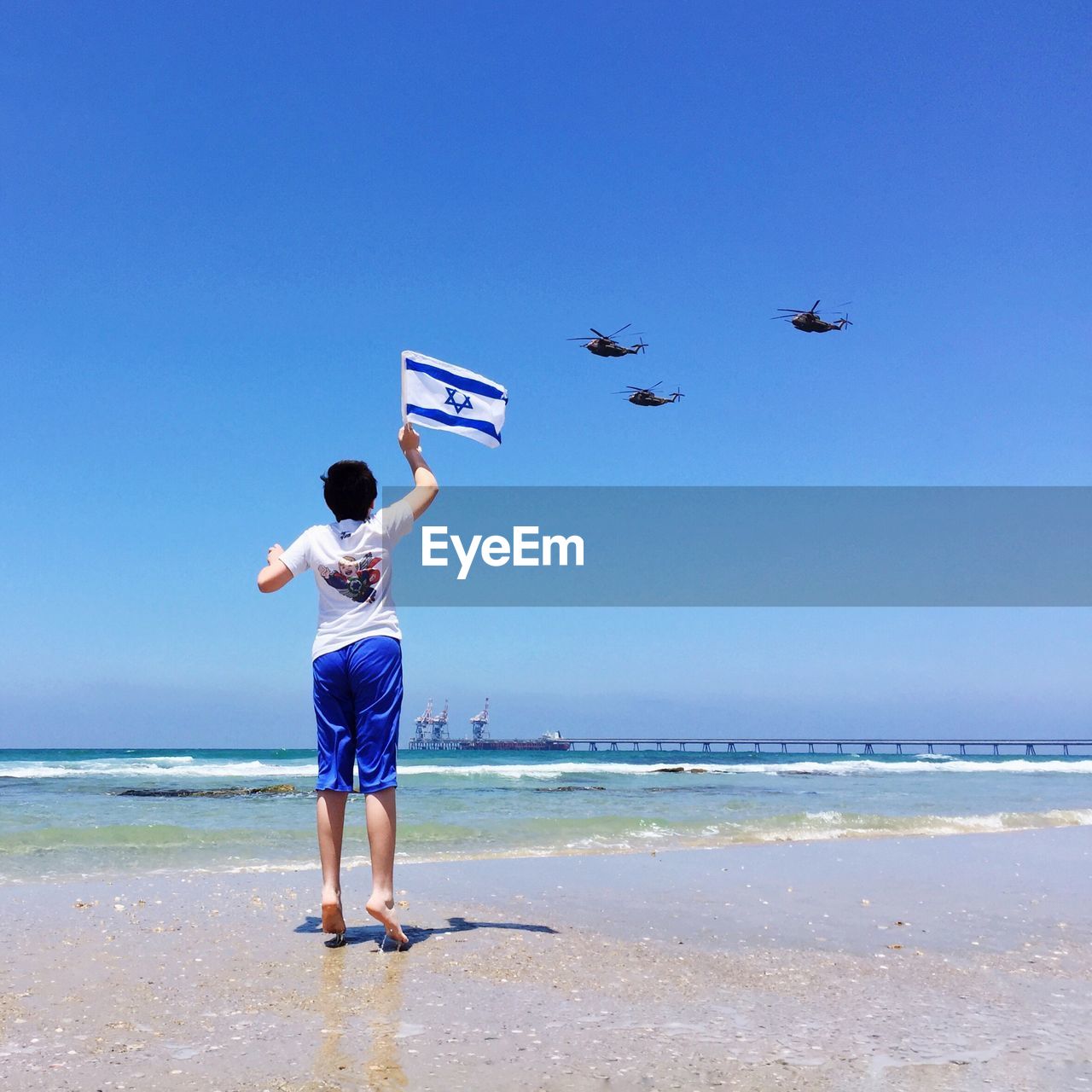 Rear view of boy jumping with israeli flag while looking at military helicopters flying over beach