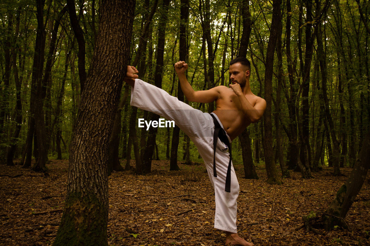 Shirtless man practicing karate against trees in forest