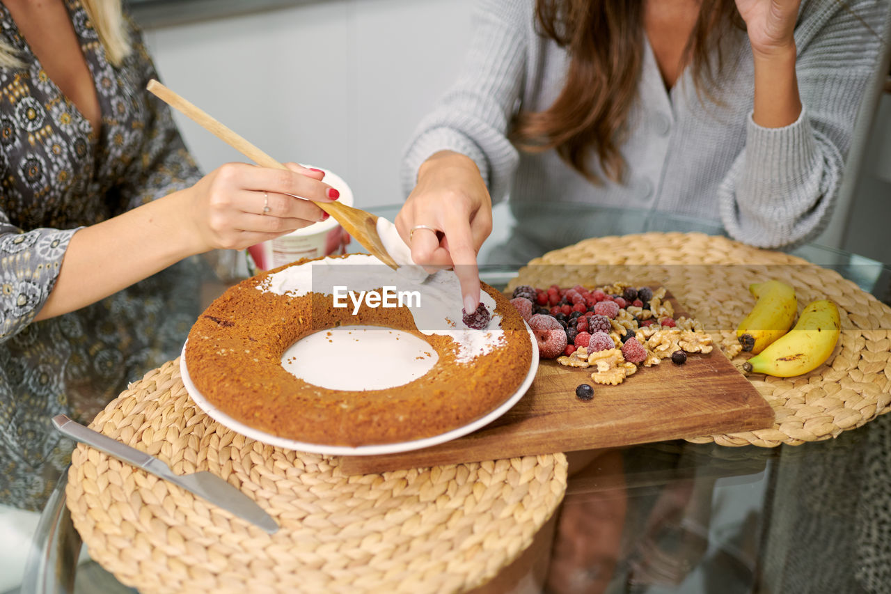 Crop anonymous female friends garnishing baked vegan banana cake with topping and fresh berries and nuts in home kitchen