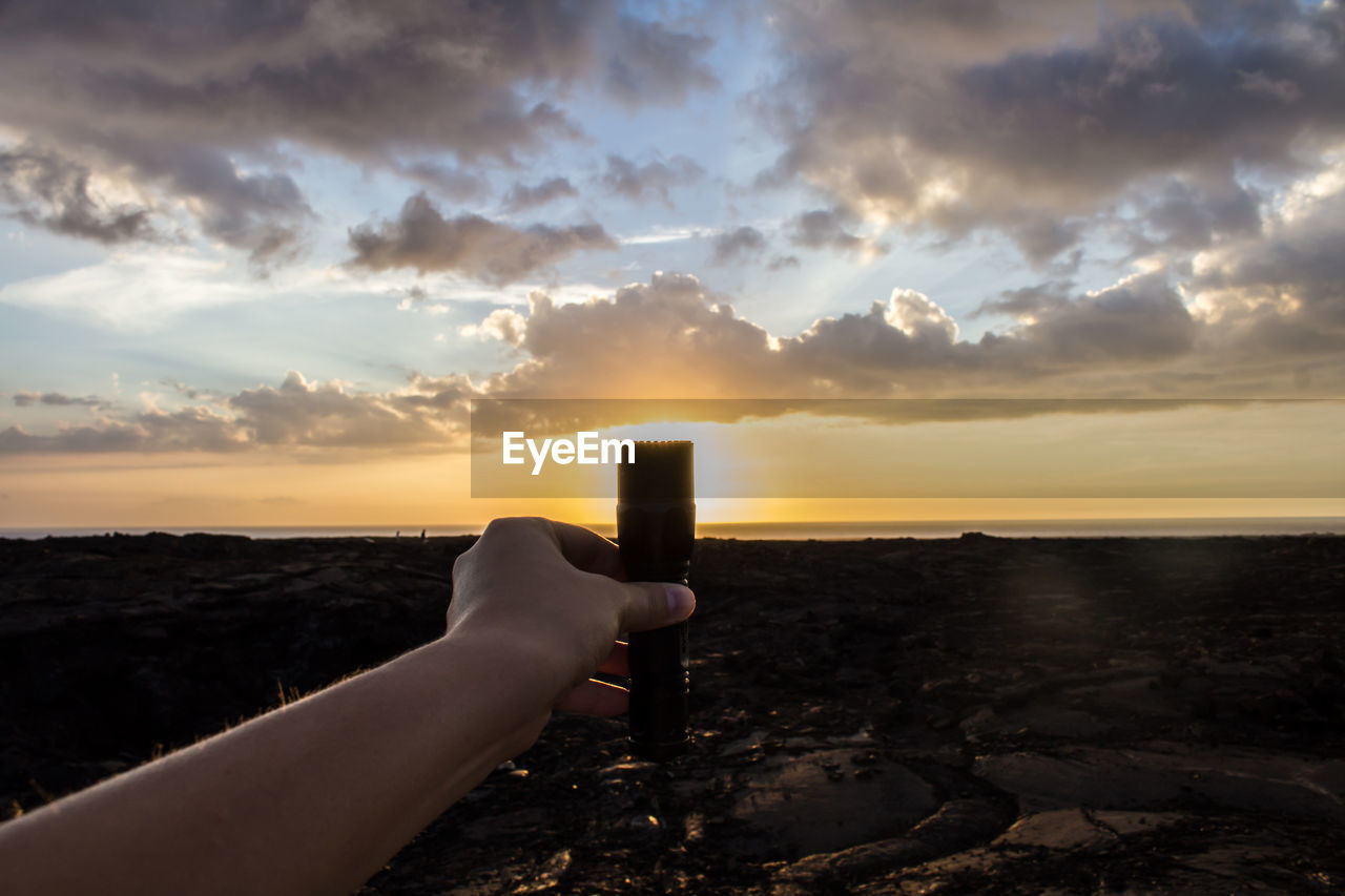 Close-up of person holding trimmer at sea shore against sunset sky