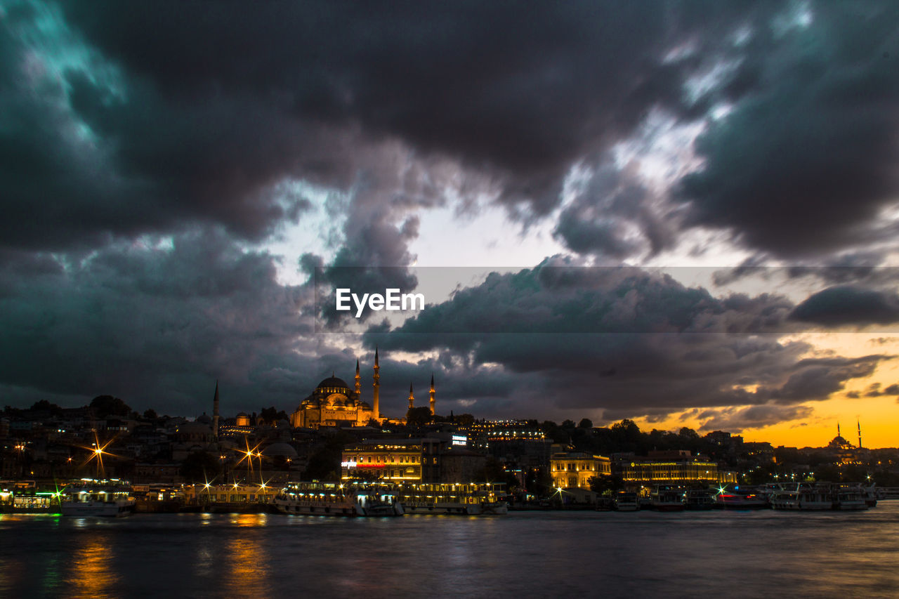 Illuminated blue mosque and city in front of sea against cloudy sky