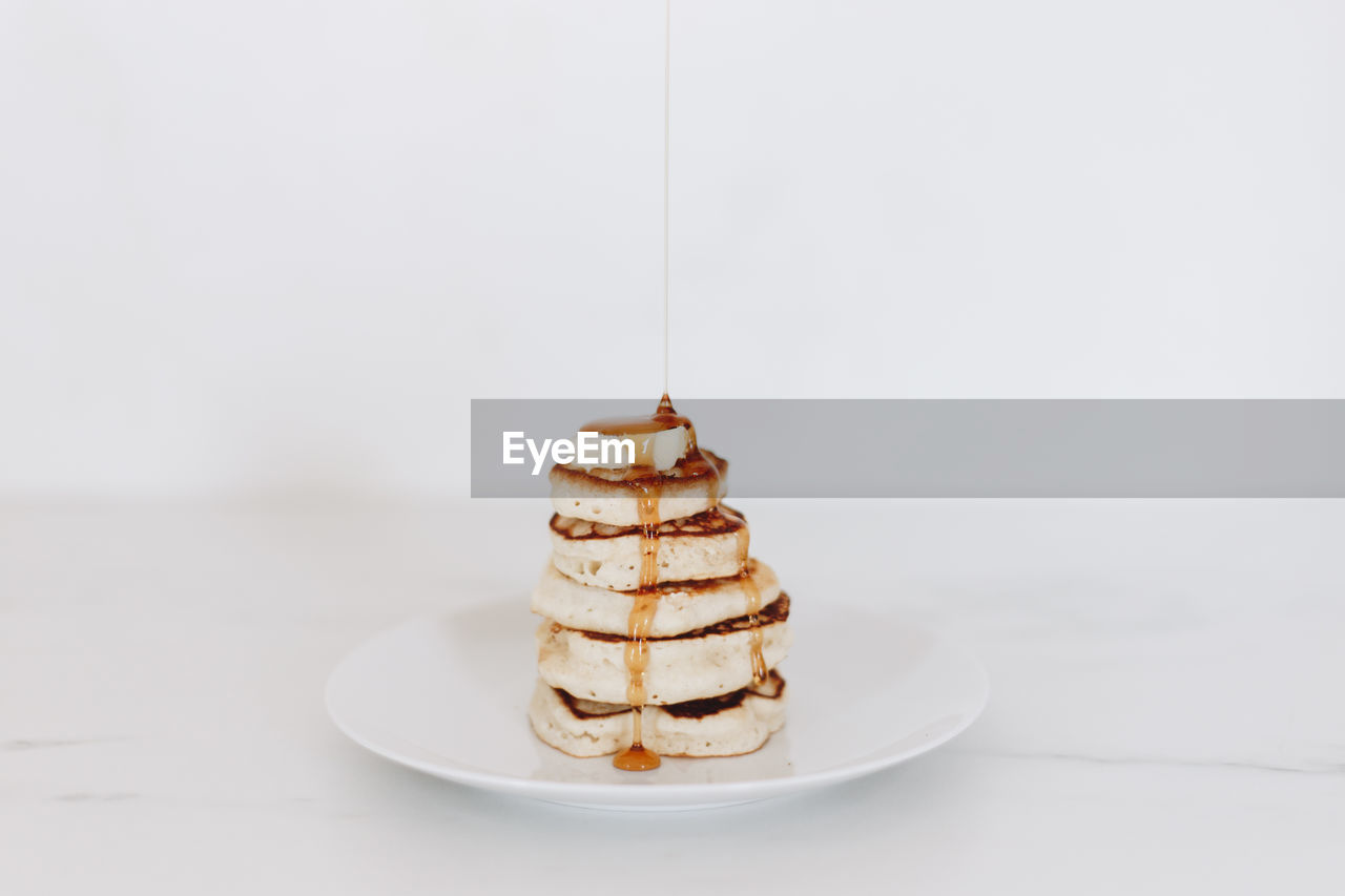 Syrup being poured on a stack of pancakes in a white kitchen scene.