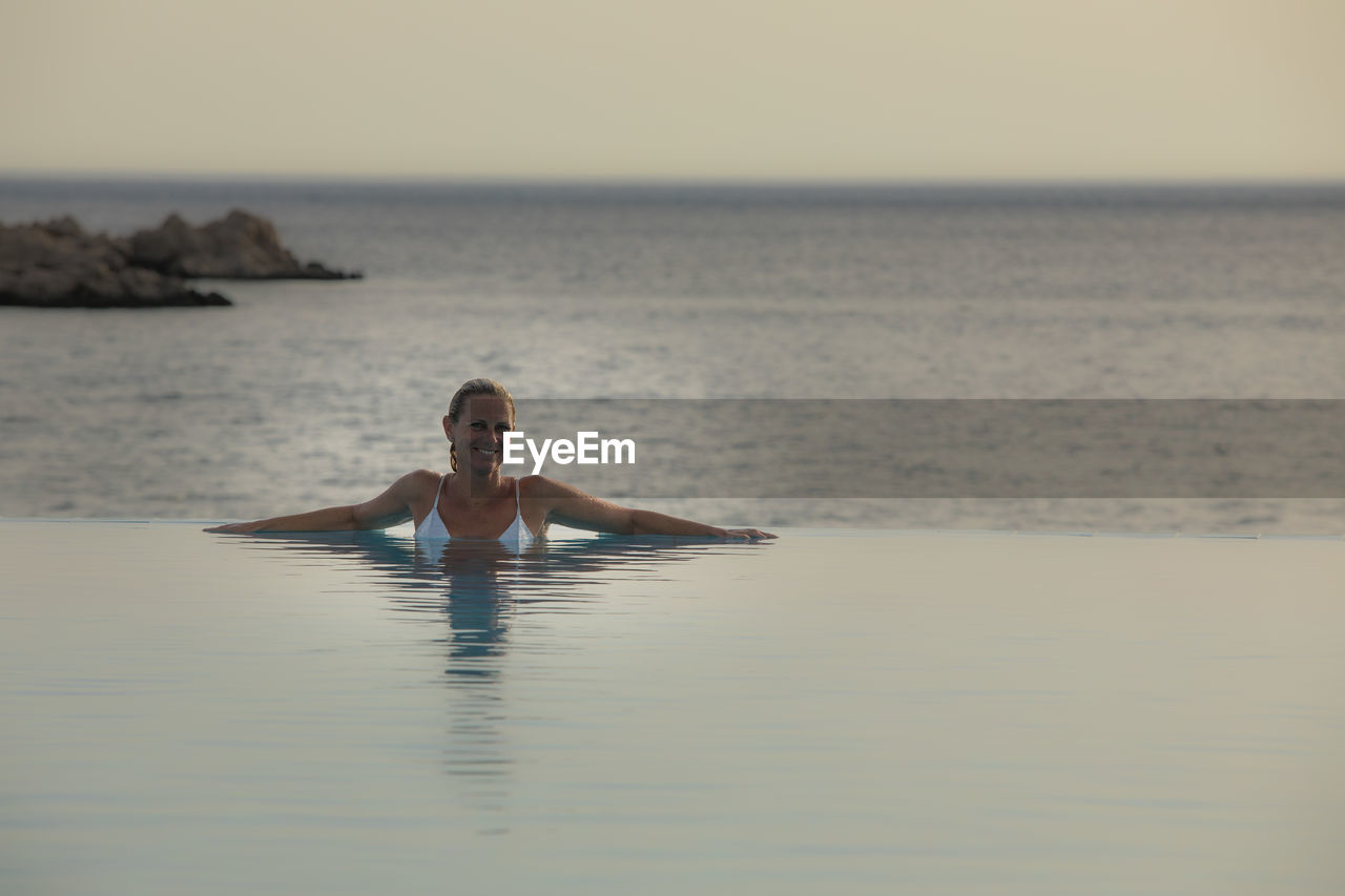 Portrait of smiling woman in infinity pool by sea during sunset