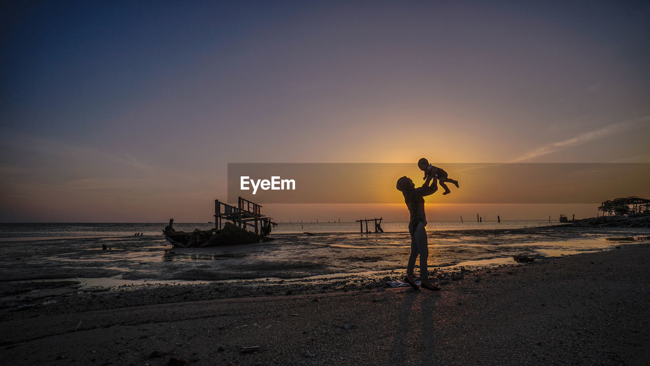 Woman holding baby while standing at beach against sky during sunset