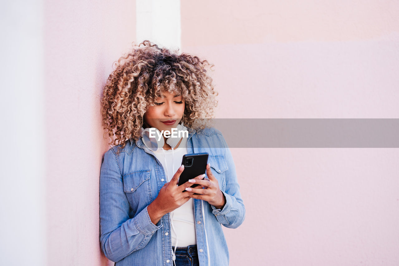 Portrait of smiling hispanic woman with afro hair in city using mobile phone and headset. lifestyle