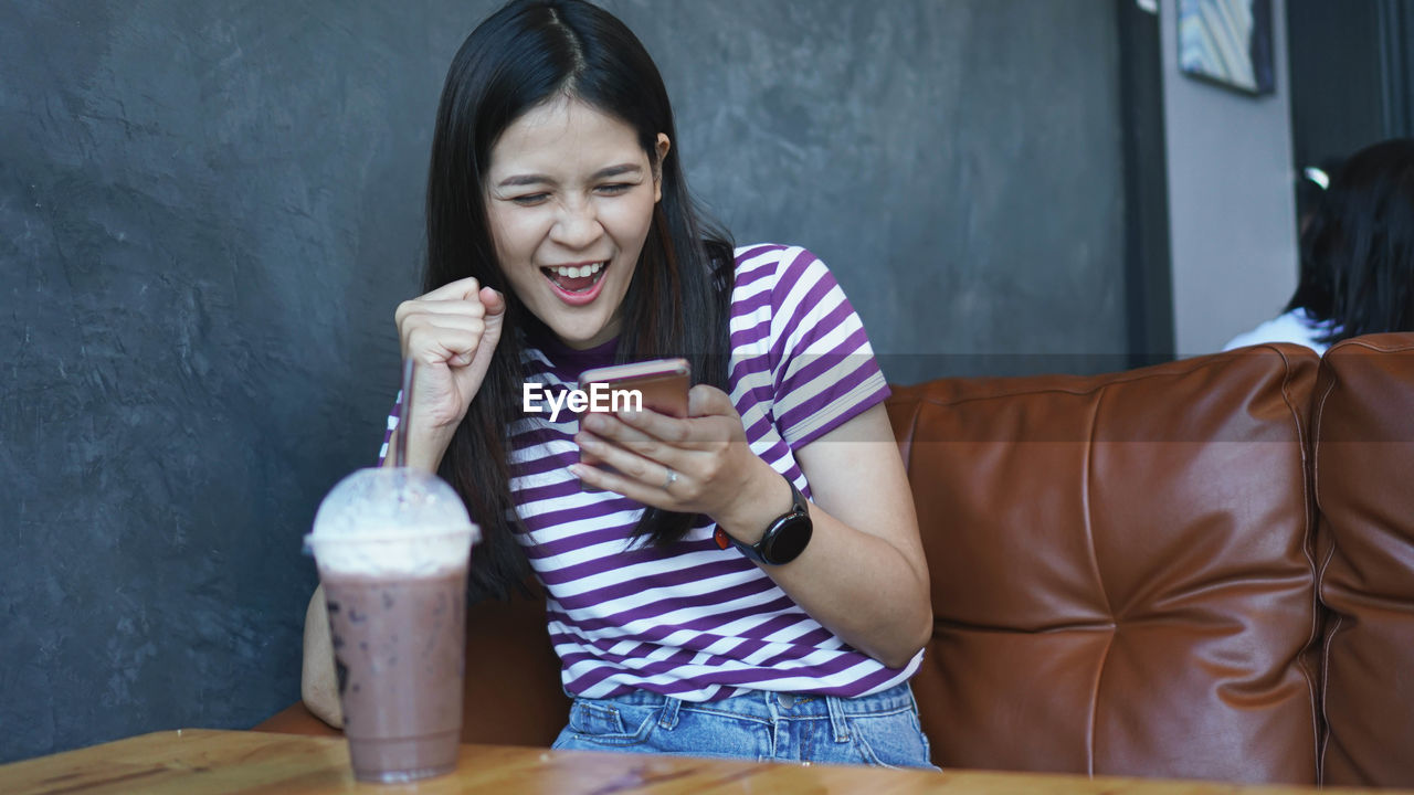 Cheerful young woman using smart phone sitting at table with coffee in cafe