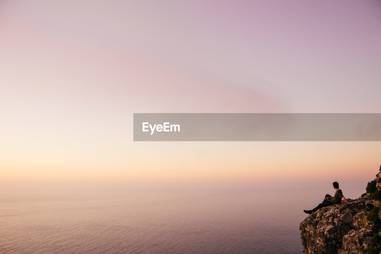 Full length of man sitting on cliff while looking at sea against sky during sunset