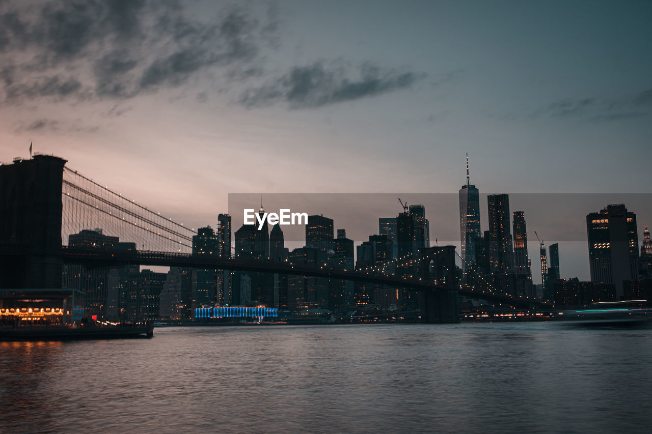 Bridge over river by buildings against sky at sunset