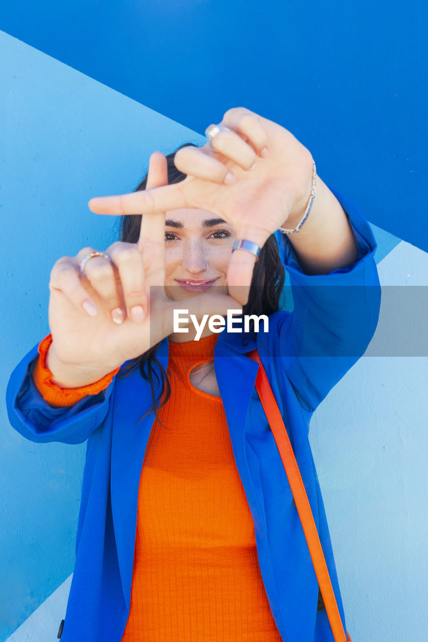 Smiling fashionable woman looking through finger frame in front of blue wall