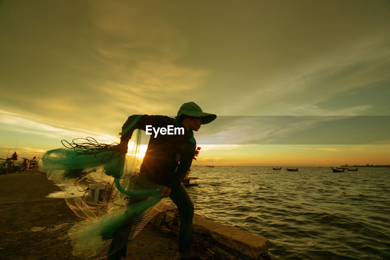 Fisherman throwing net in sea against sky during sunset