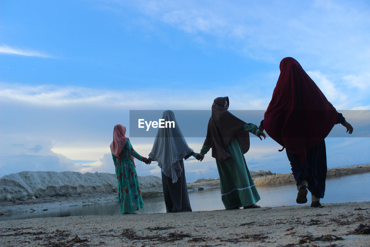 Rear view of women holding hands while standing at beach against sky