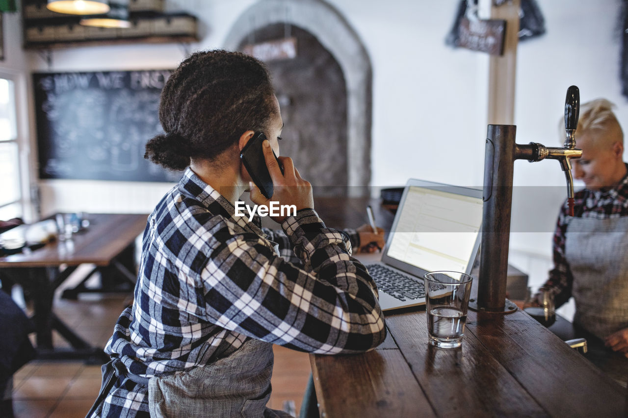 Female manager talking on mobile phone while using laptop at bar counter