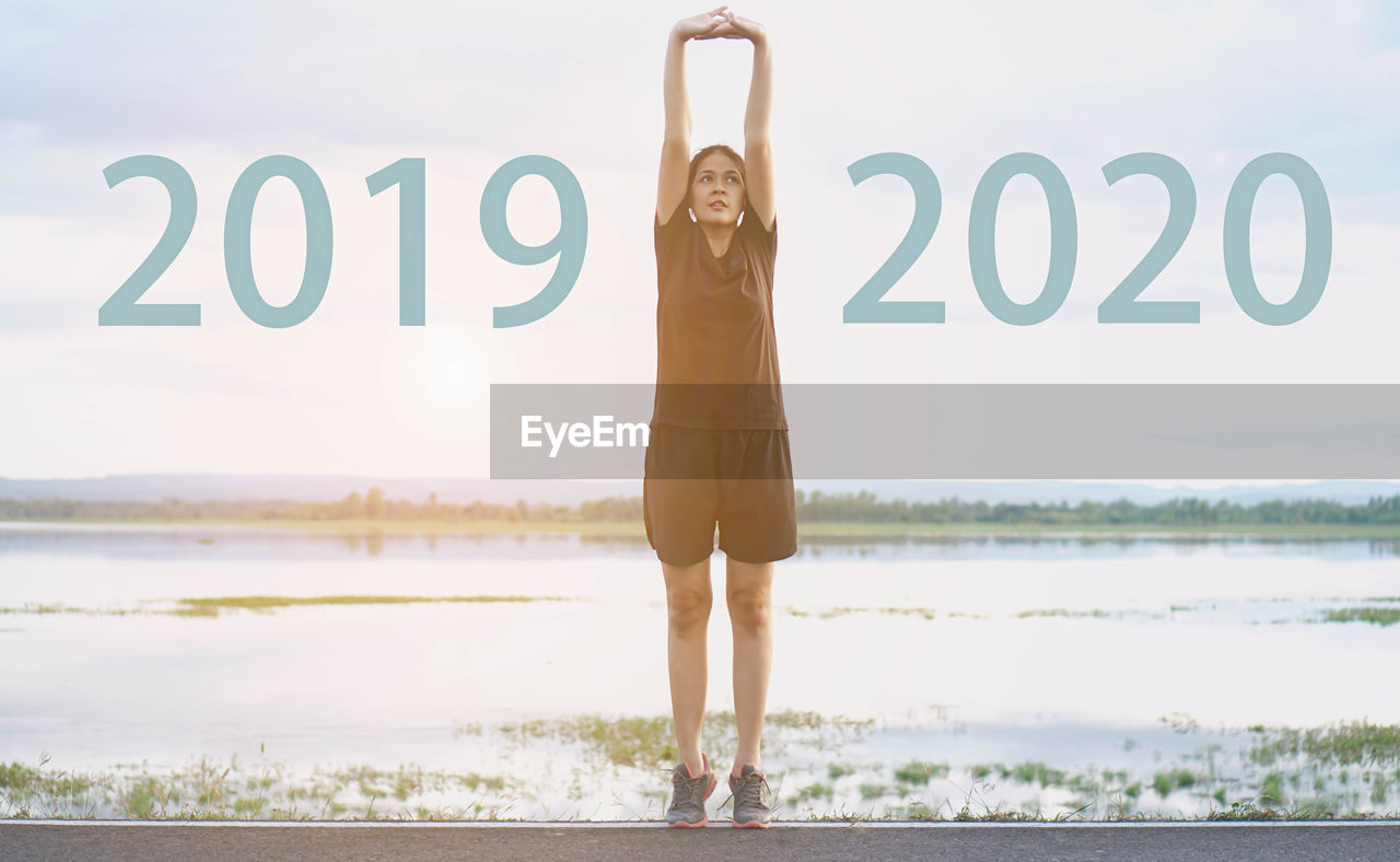Digital composite image of young woman stretching on road by 2020 and 2019 against lake and sky