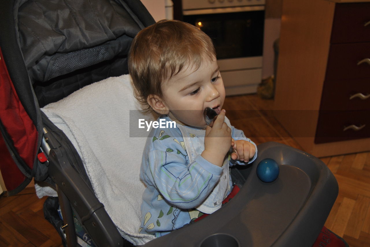 Cute boy licking spoon while sitting on baby stroller at home