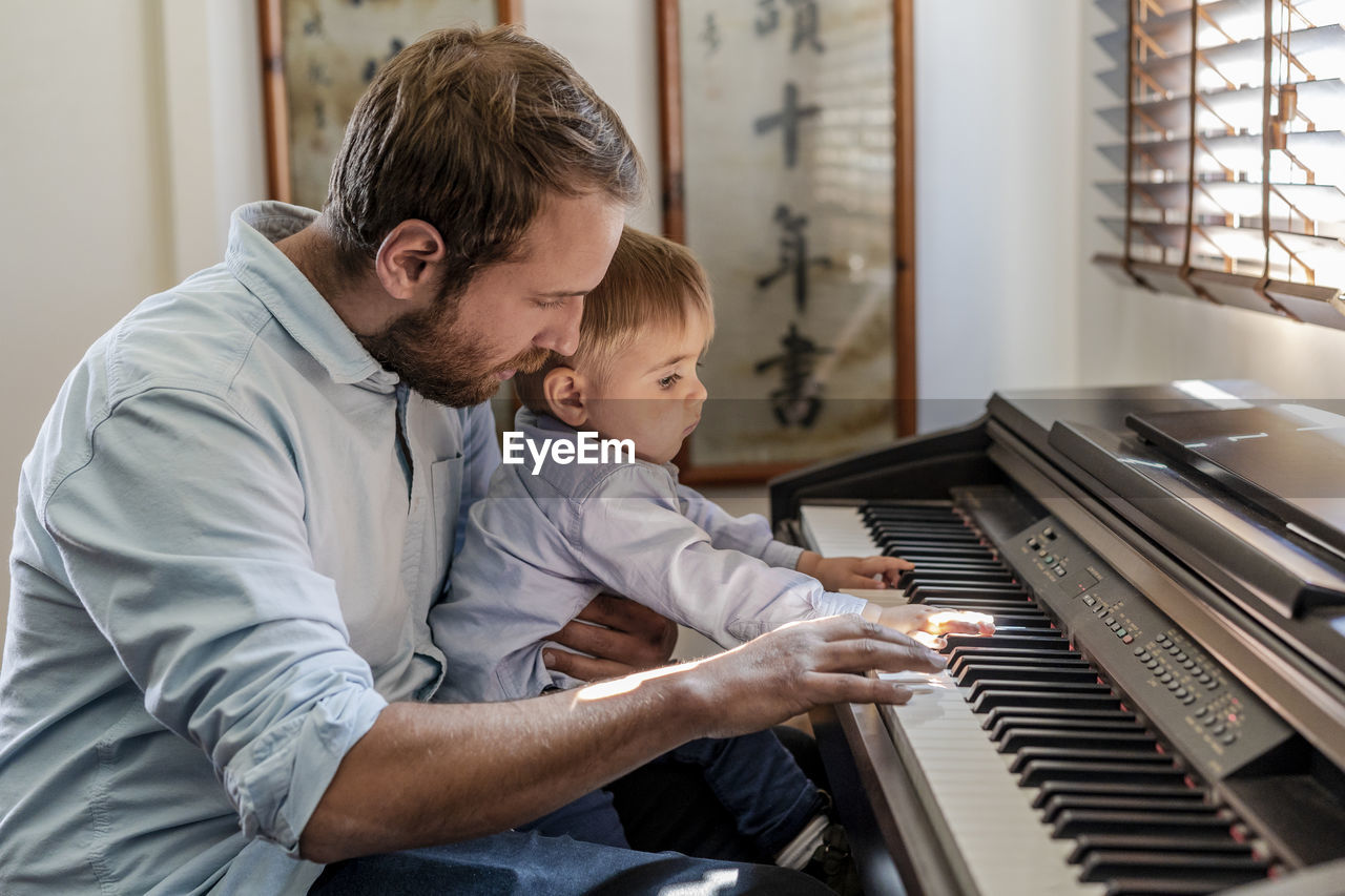 Father teaching piano to son sitting on lap at home