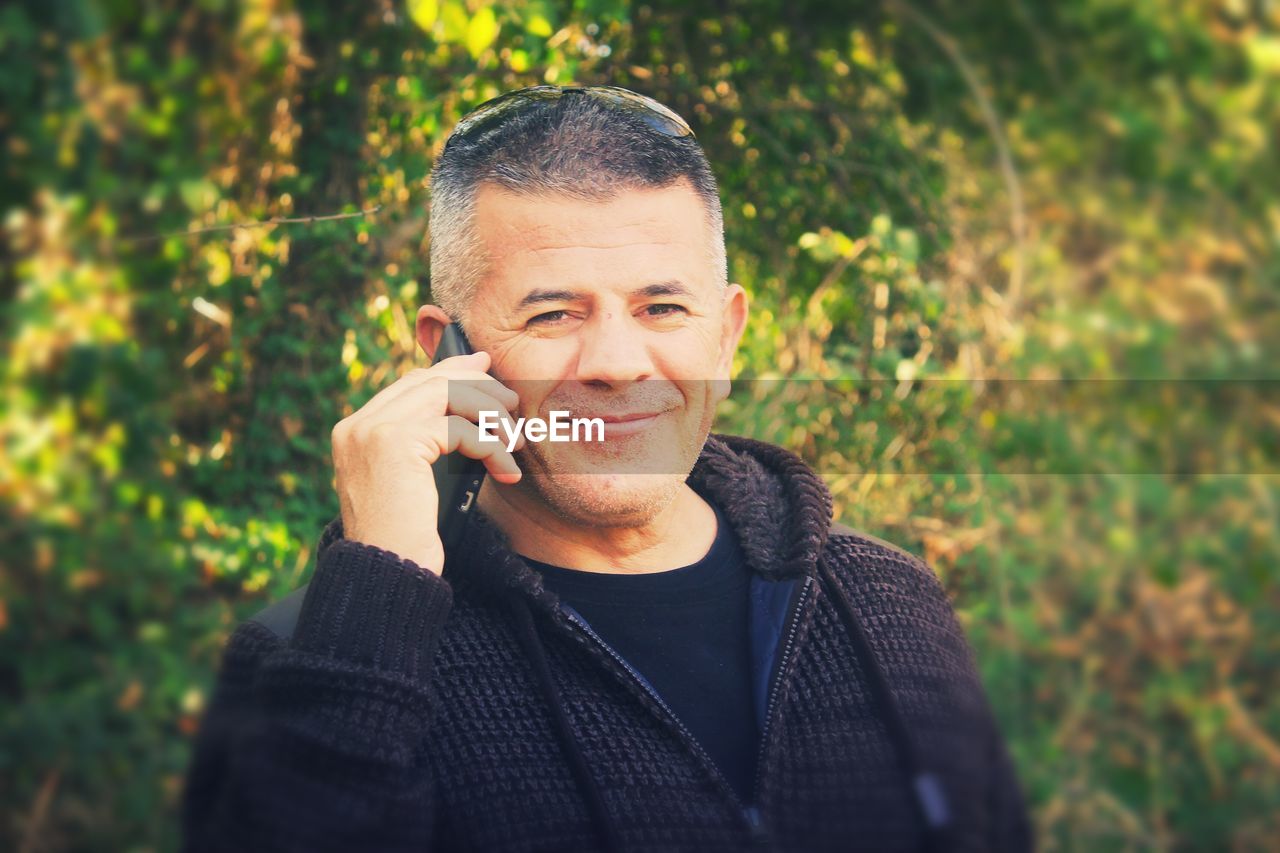 Portrait of smiling mature man talking on mobile phone while standing against plants