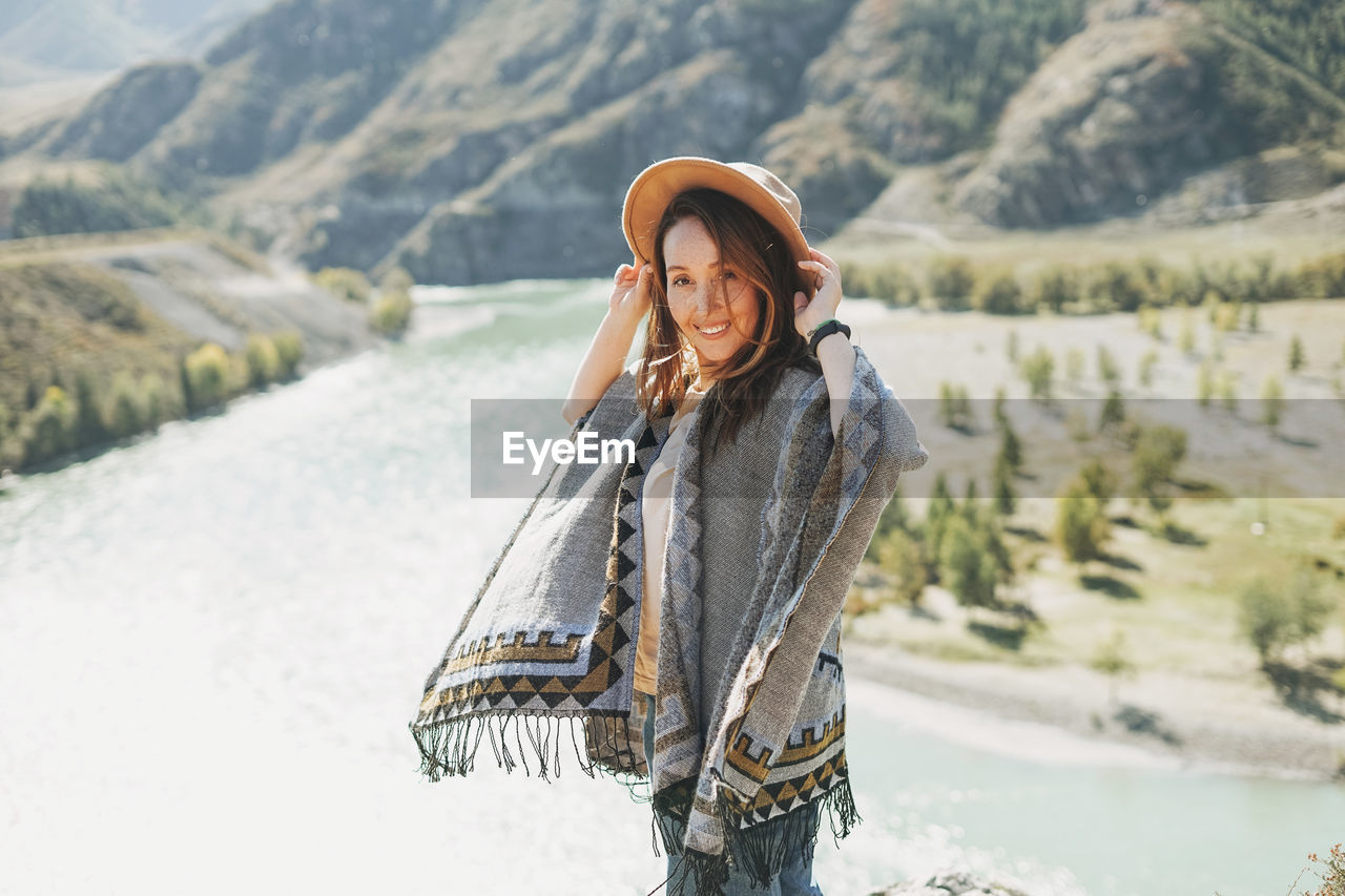Carefree brunette young woman traveler in poncho from back on background of mountain river