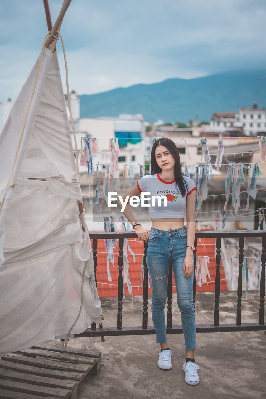 Full length portrait of young woman standing by tent against cloudy sky in city