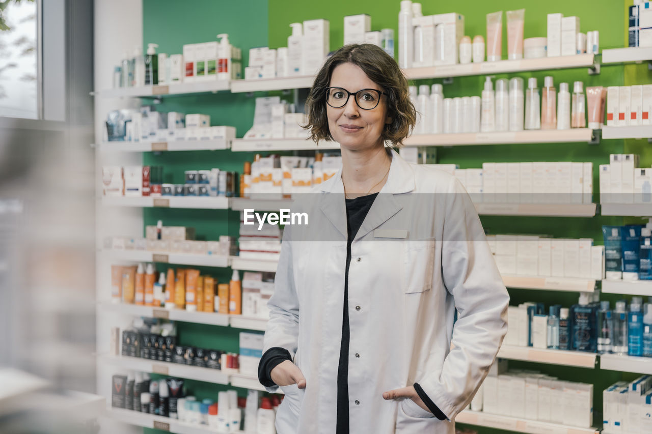 Confident pharmacist with hands in pockets standing in chemist shop