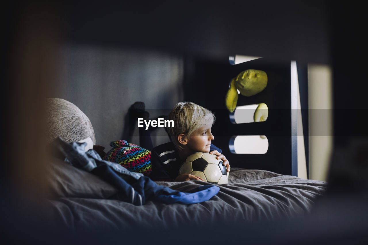 Boy with soccer ball looking away while lying on bed