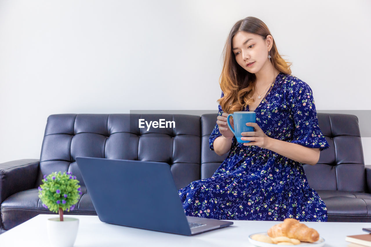 YOUNG WOMAN USING PHONE WHILE SITTING ON TABLE INDOORS